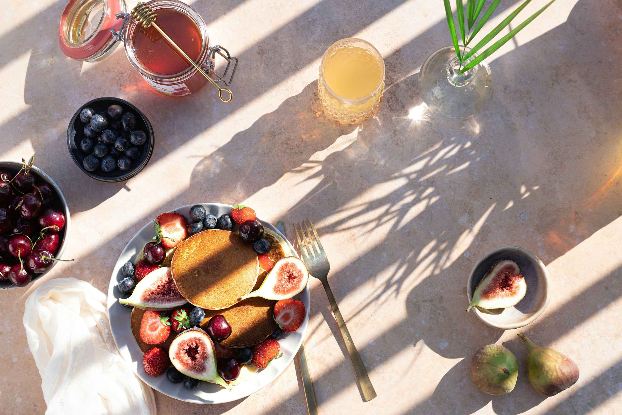 Colazione vista dall’alto su un piano chiaro, nel sole: pancake con fichi, fragole e mirtilli, una ciotola di ciliegie, miele e un succo, fra le ombre nette di una pianta.