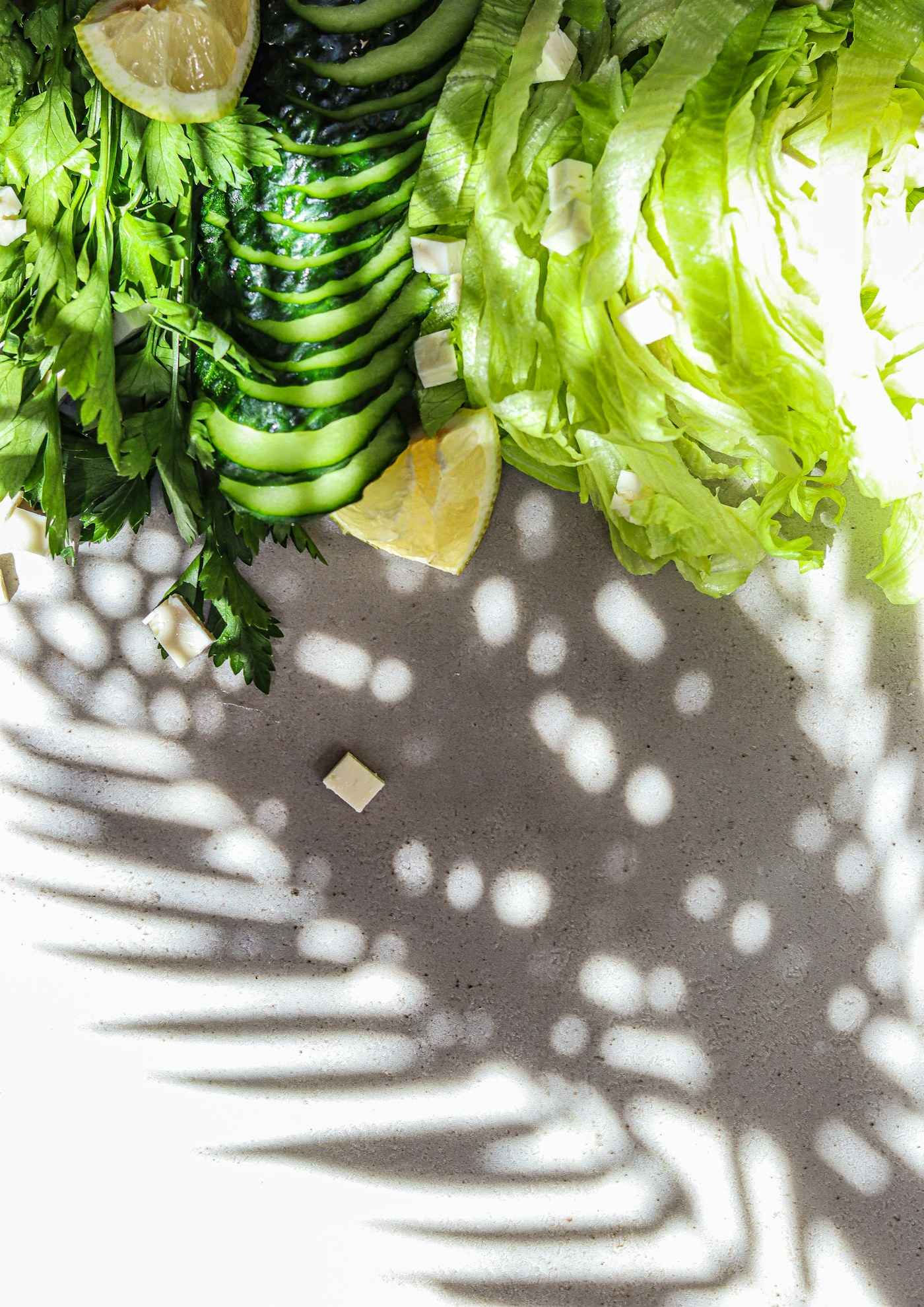 Lattuga, cetriolo a fette, prezzemolo, spicchi di limone e cubetti di formaggio visti dall’alto su un piano chiaro, sotto l’ombra a macchie di una palma.